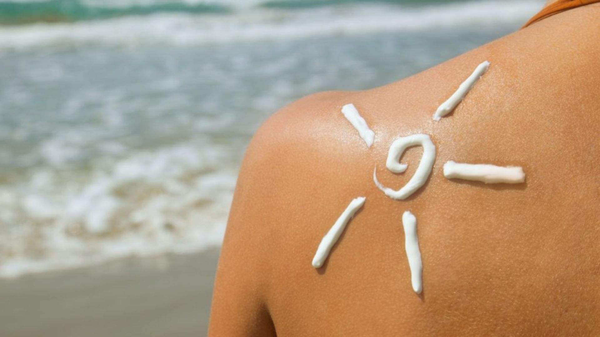 A woman applies sun cream to her back while relaxing on the beach under a clear blue sky.