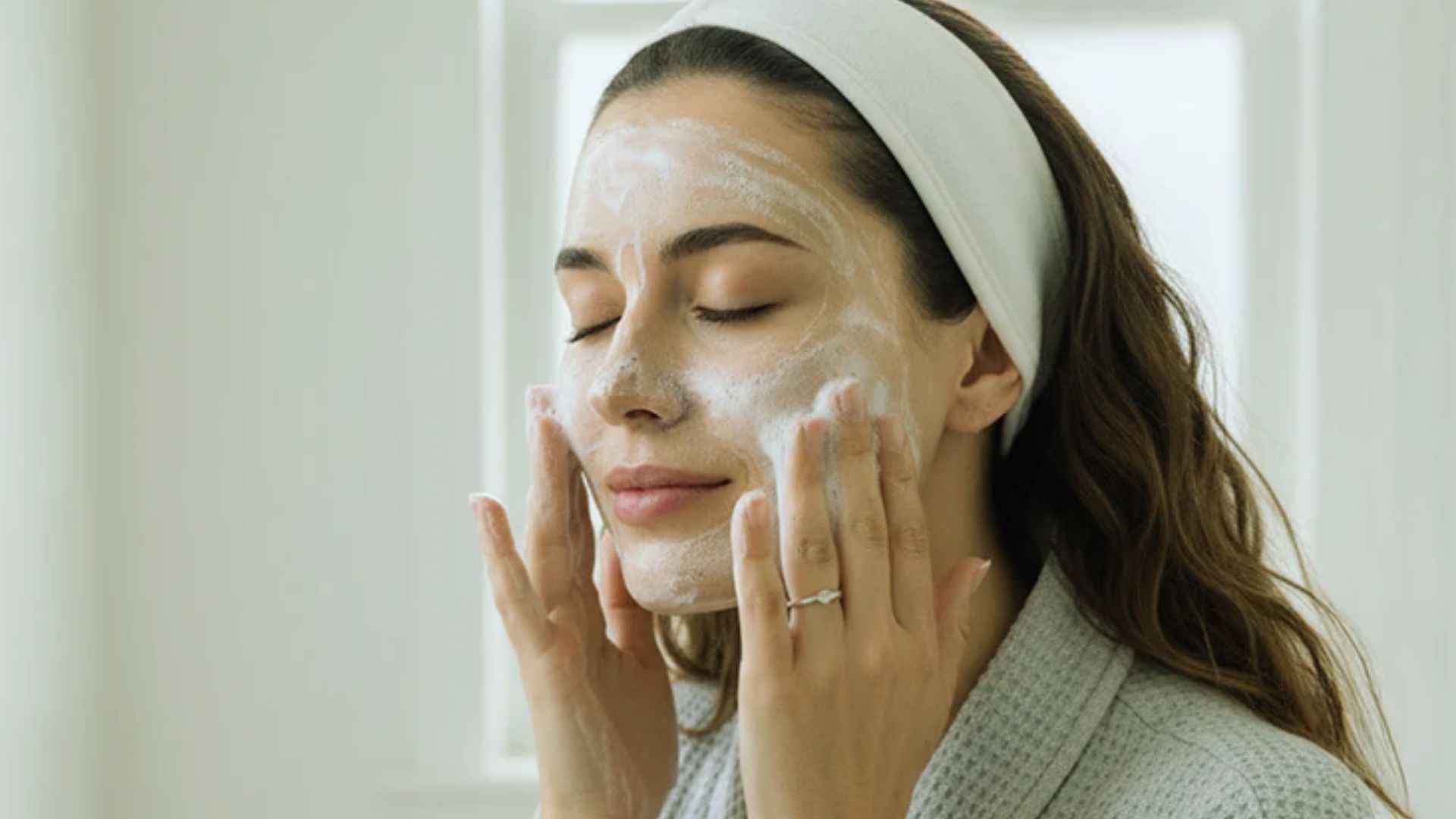 A woman applies a facial mask to her face, focusing on skincare and self-care in a bright, well-lit room.