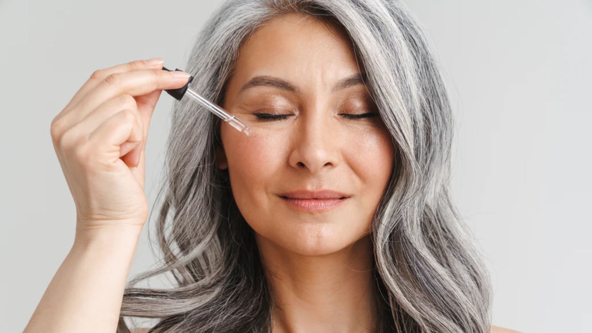 A woman with gray hair holds a brush, preparing to style her hair with a focused expression.