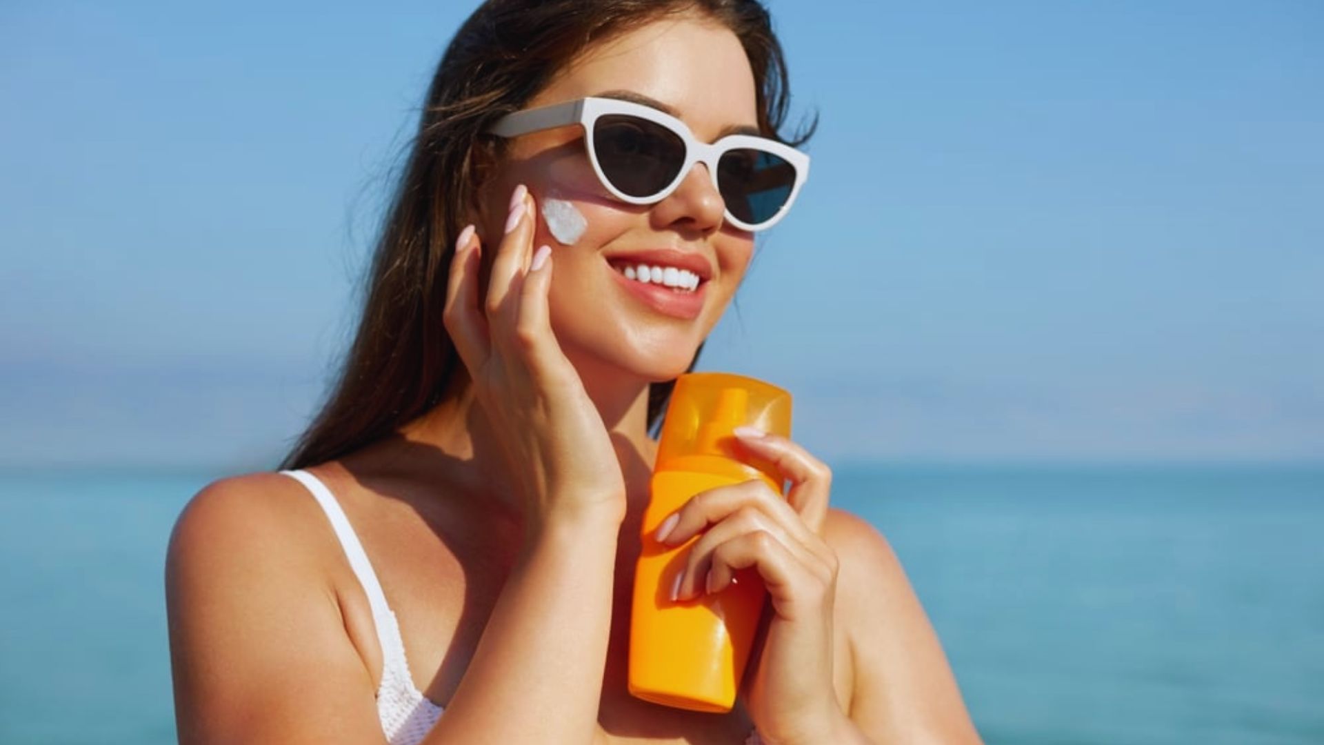 A woman wearing sunglasses and a white shirt holds an orange sunscreen bottle, ready for sun protection.