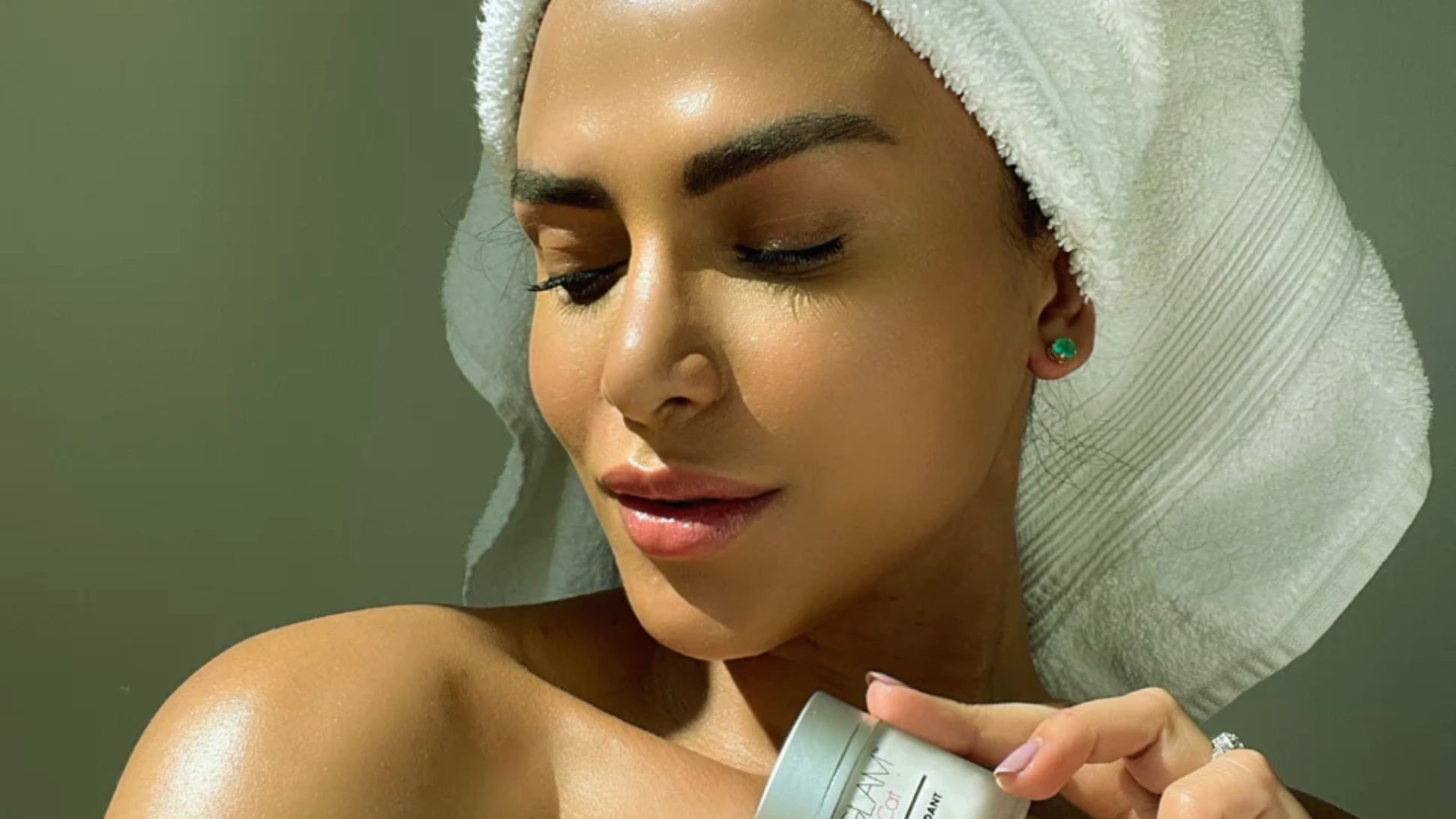A woman wrapped in a towel holds a jar of cream, preparing for her skincare routine.