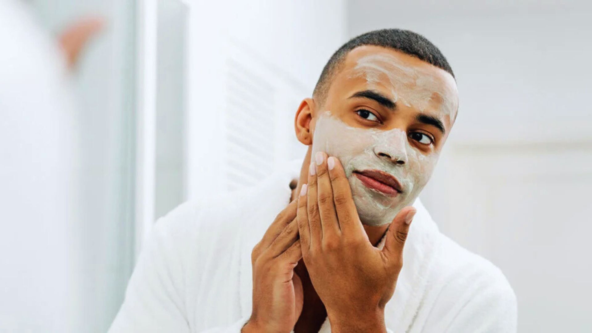 A man in a bathrobe applies a facial mask, focusing on his skincare routine in a relaxed setting.