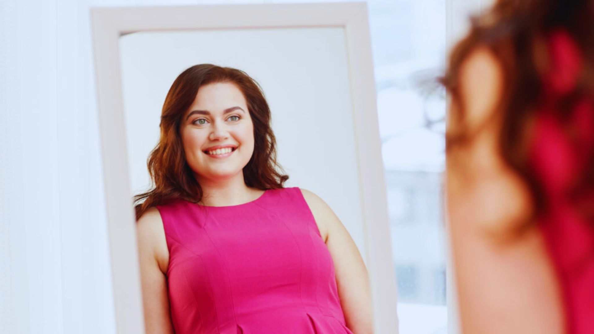 A woman in a pink dress gazes at her reflection in a mirror, admiring her appearance.