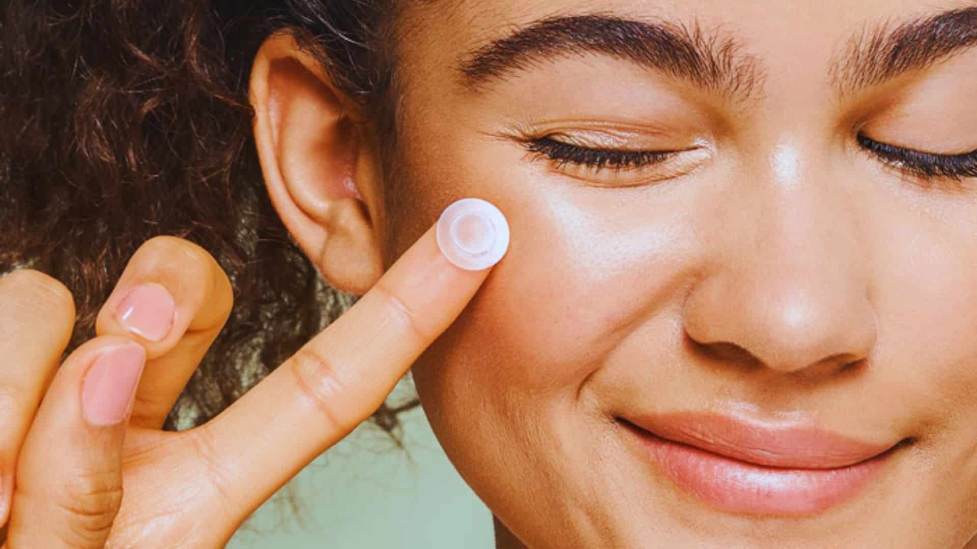 A woman applies cream to her face, focusing on skincare in a bright, well-lit setting.
