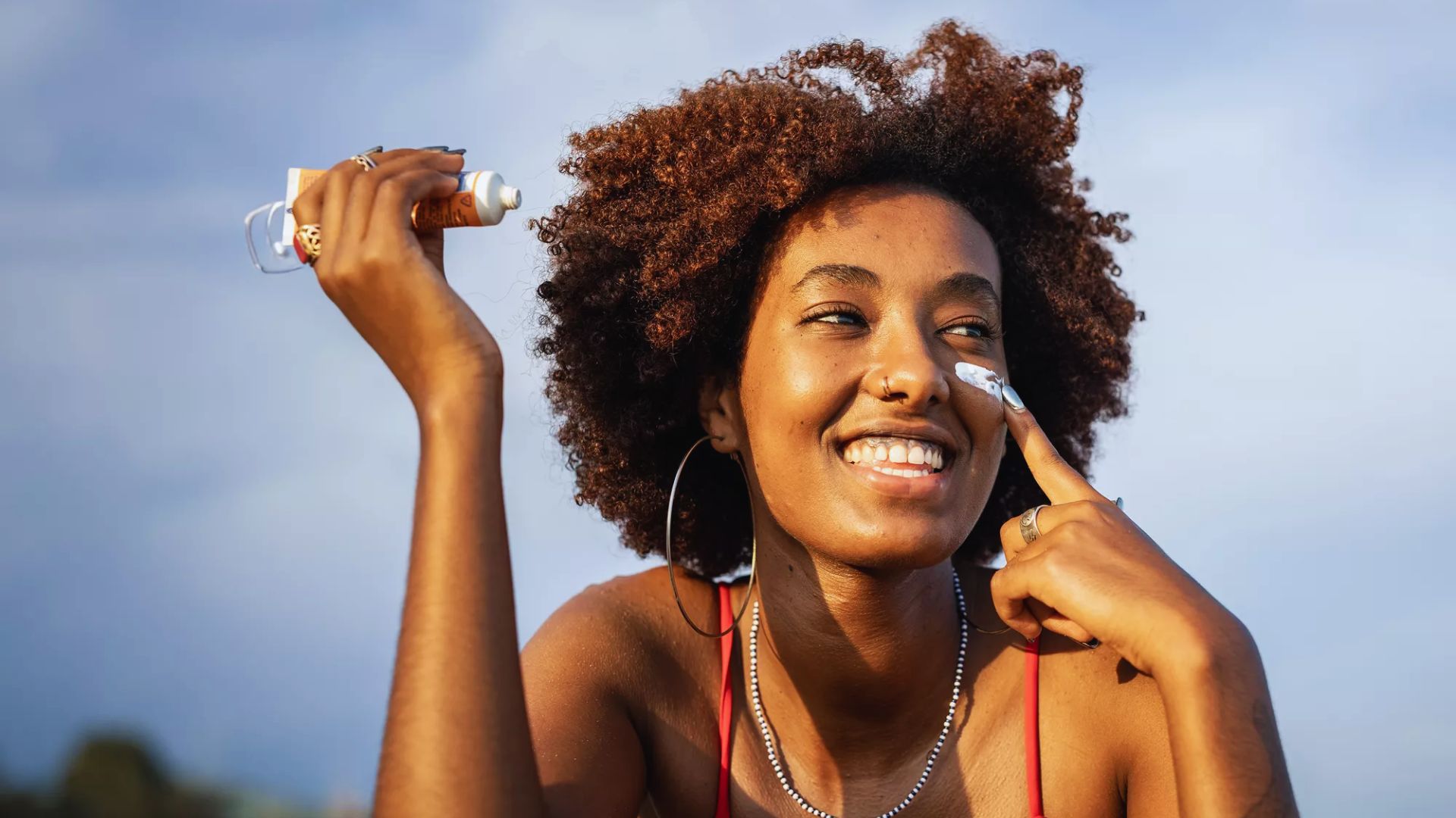 A smiling woman with an afro hairstyle holds a bottle of sunscreen, promoting sun safety and skincare.