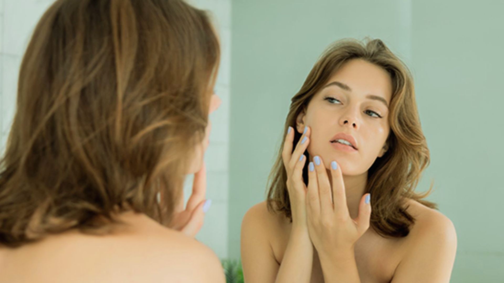 A woman applies skin care products while gazing into a mirror, focusing on her reflection and skincare routine.