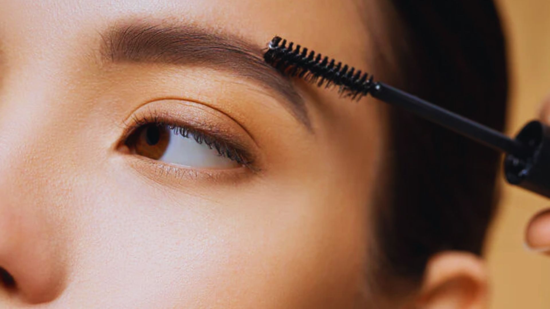 A woman applies mascara to her eyebrows using a brush, enhancing her facial features with precision.