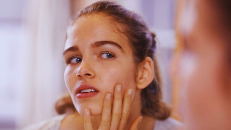 A woman gazes into a mirror, gently touching her face with a thoughtful expression.