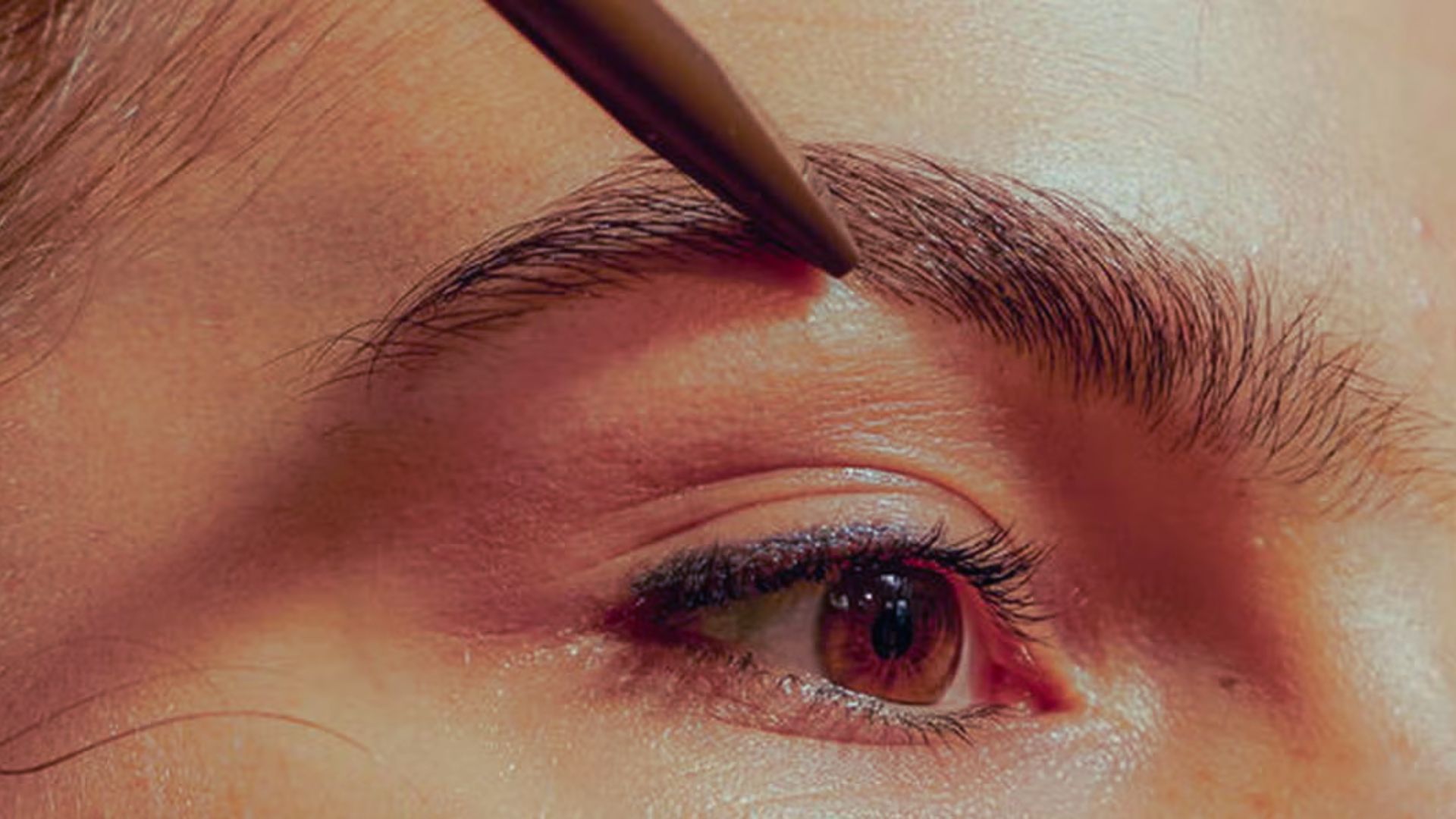 A woman applies eyebrow makeup with a brush, focusing on enhancing her brows in a well-lit setting.