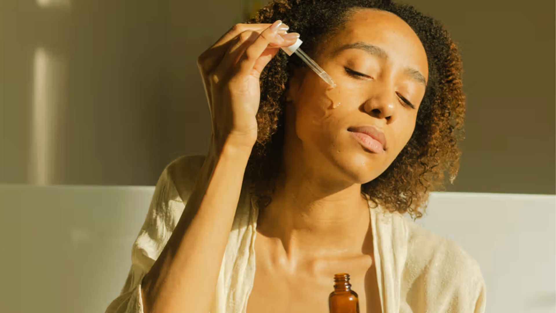 A woman is seen applying serum to her face, emphasizing her skincare regimen in a clean, well-lit environment.