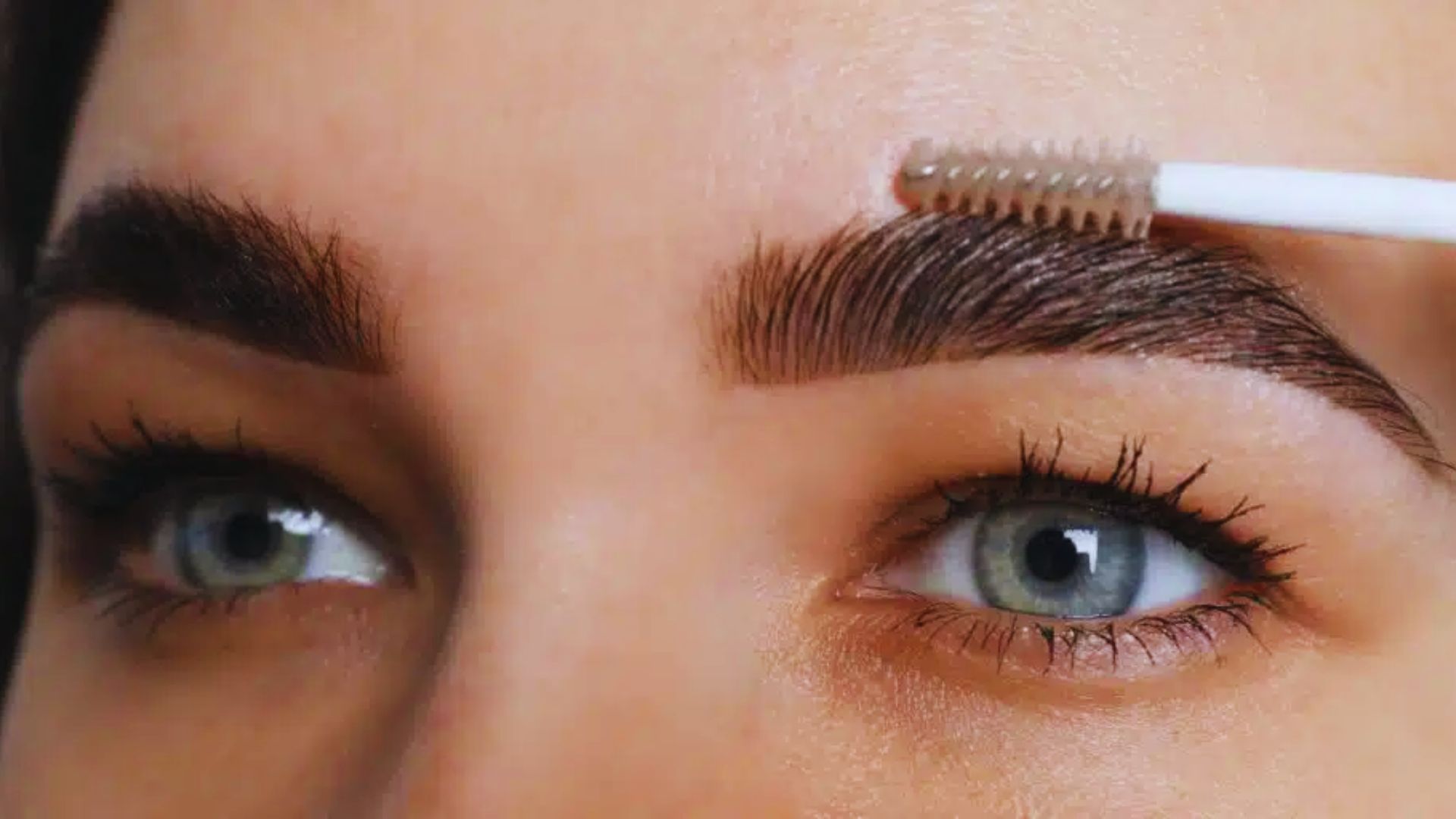 A woman applies eyebrow makeup with a brush, focusing intently on her reflection in a mirror.