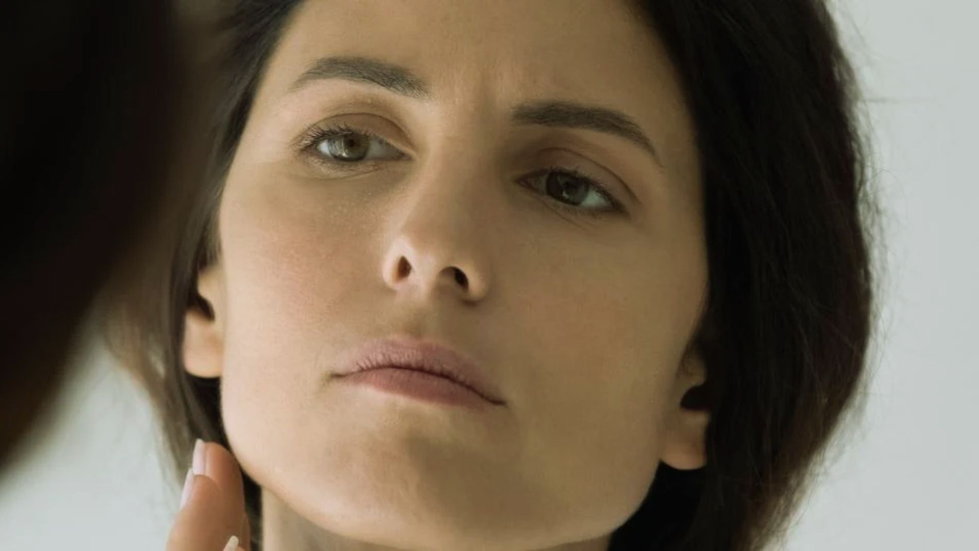 A woman with a contemplative expression looks in the mirror, applying skincare with her hand. The light setting is neutral, conveying a calm mood.