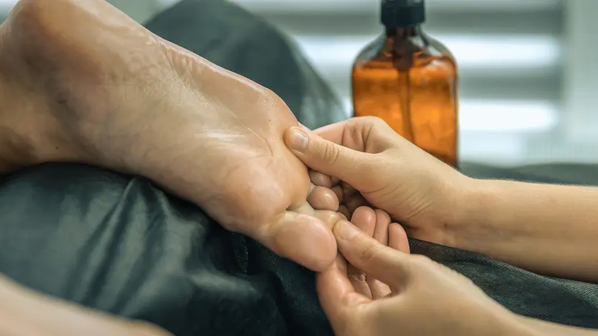 Hands gently massaging the sole of a foot on a dark cloth surface, with a brown glass bottle in the background, conveying relaxation and care.