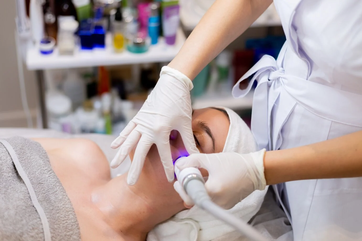 A woman receiving a laser facial treatment, lying on a treatment bed with a professional applying the device to her face.