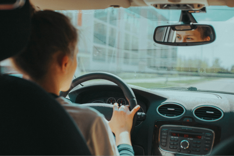 A person driving a car, focusing on the road. The rearview mirror shows their eyes. Daylight streams in, creating a calm and attentive atmosphere.