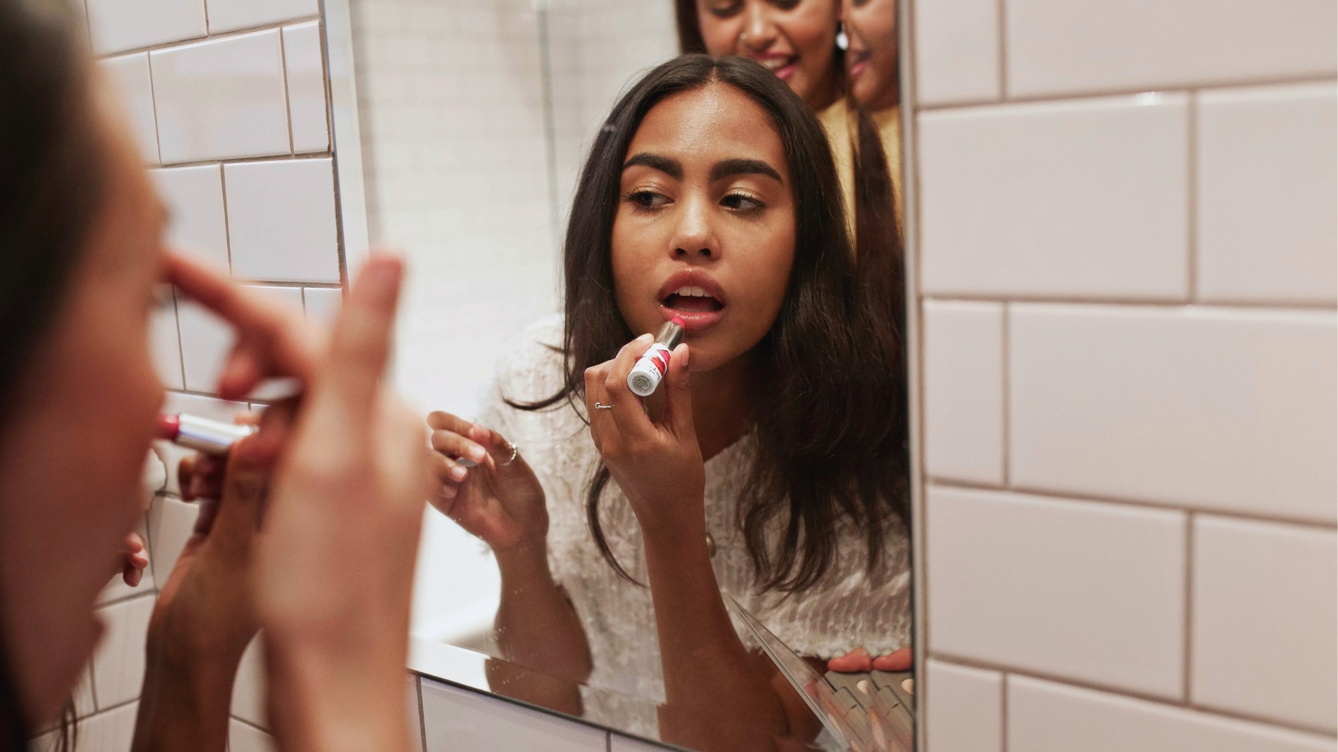 A woman is applying lip gloss while looking into a tiled bathroom mirror. Several friends in the background smile, creating a fun and lively atmosphere.