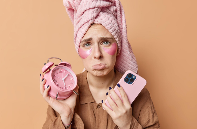 A woman wearing a pink towel on her head is holding a pink alarm clock and a pink phone, looking at the camera.
