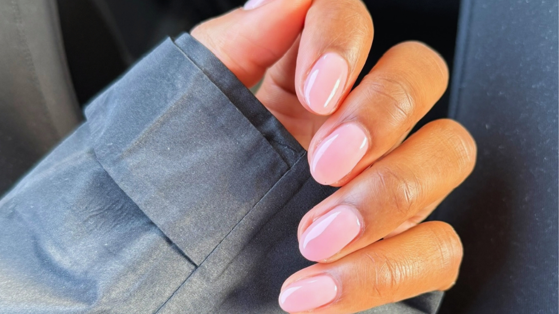 A hand with neatly manicured nails painted in glossy light pink is shown resting on a dark gray fabric. The image conveys a sense of elegance and care.