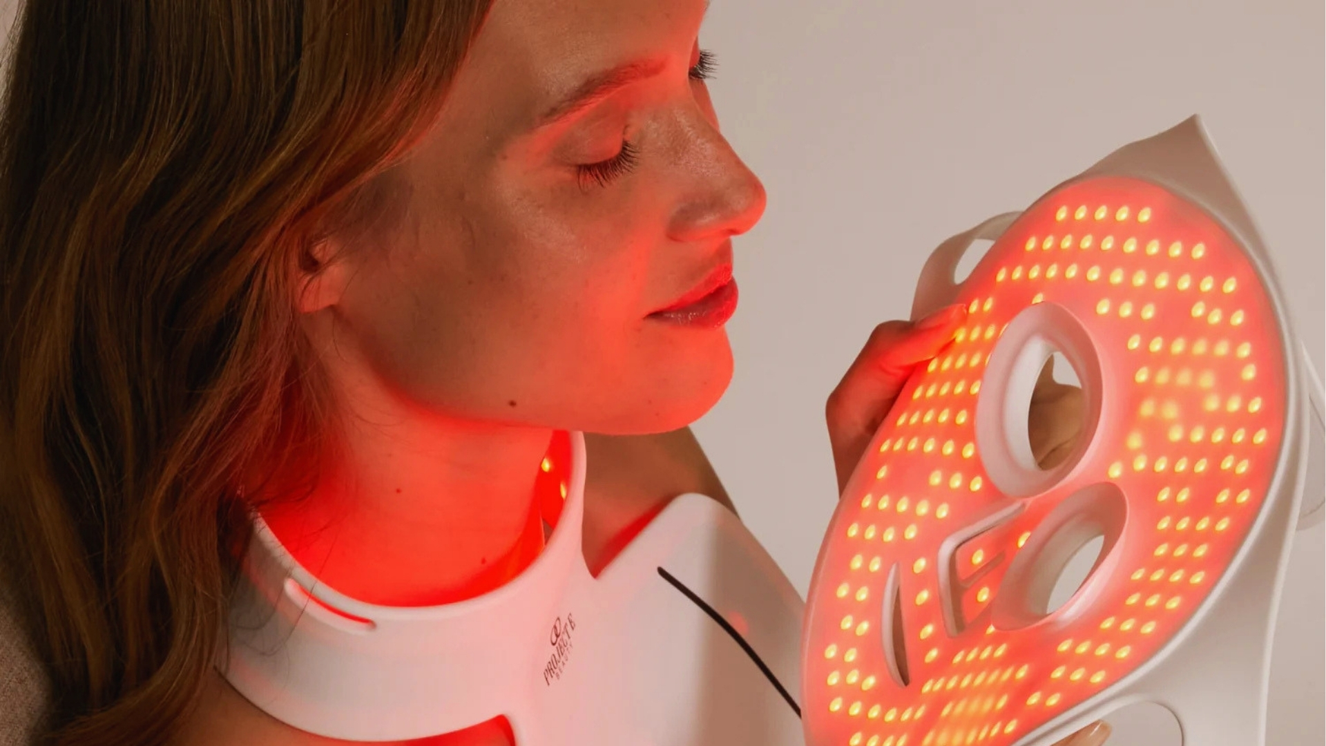 A woman gently holds a glowing LED facial mask emitting red light therapy. She seems relaxed and content, suggesting a soothing skincare routine.