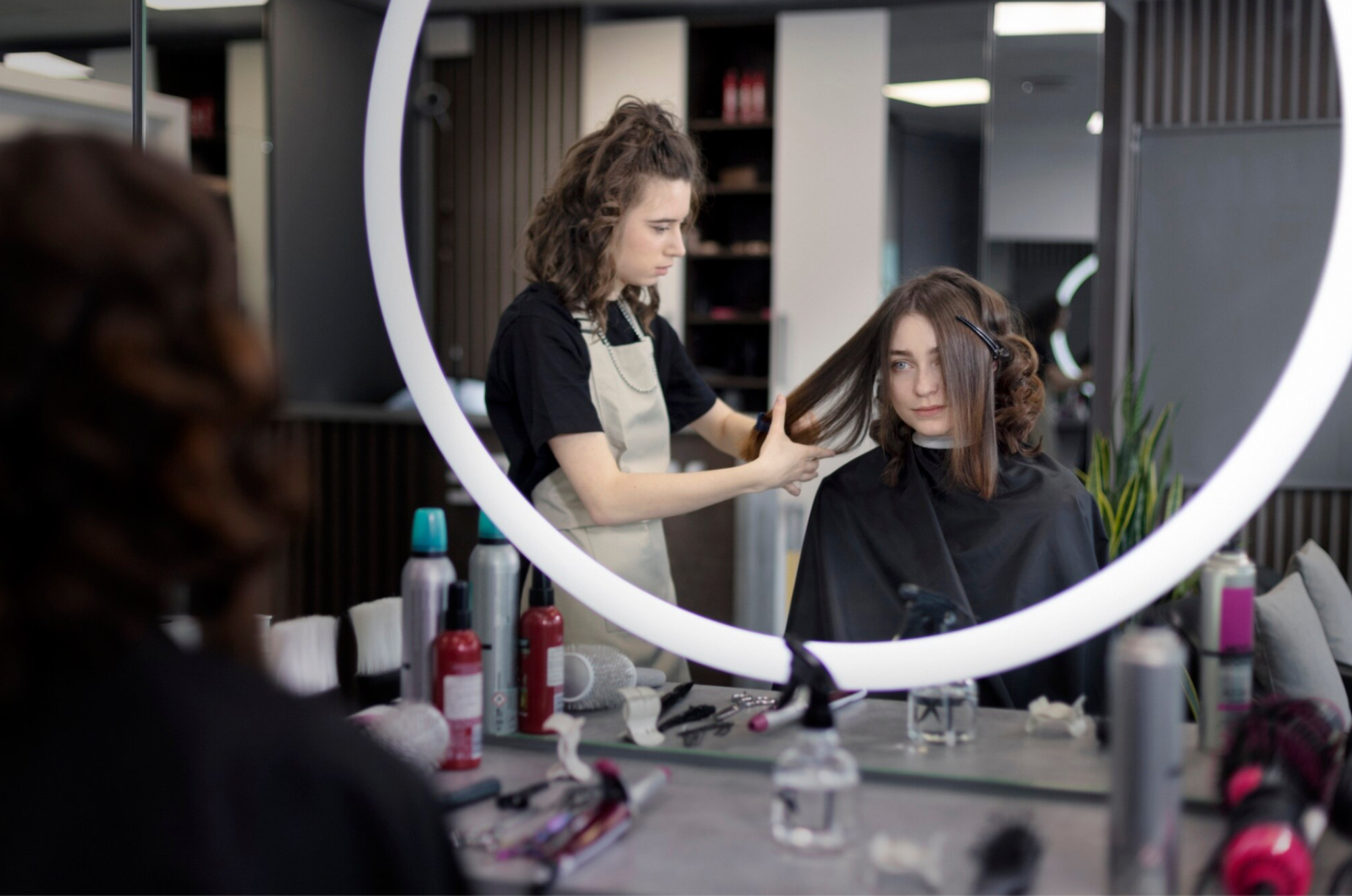 A woman sits in a salon chair as a stylist cuts her hair.