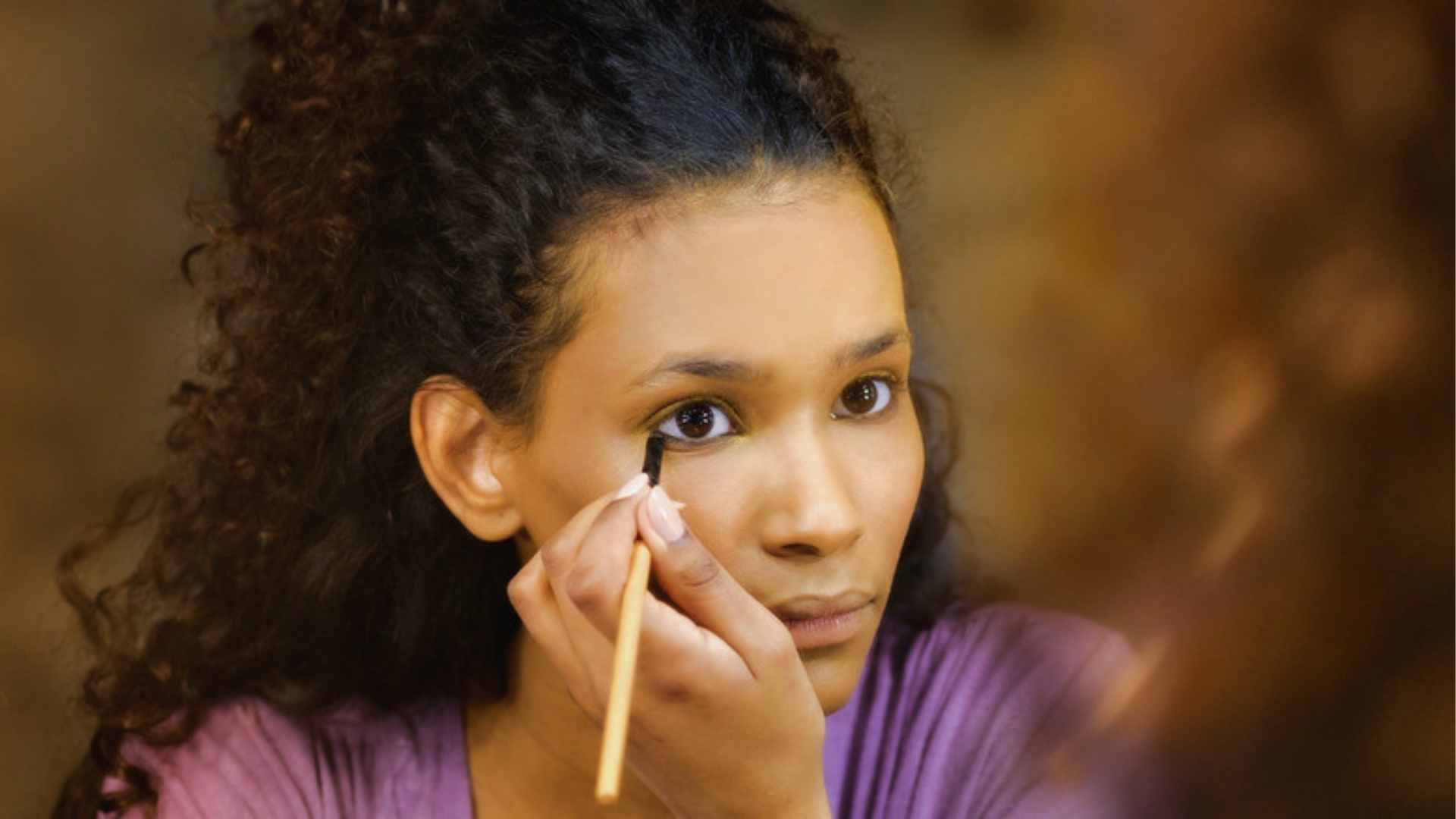 A person with curly hair intently applies black eyeliner to their lower eyelid using a pencil, reflected in a mirror. The scene conveys focus and precision.