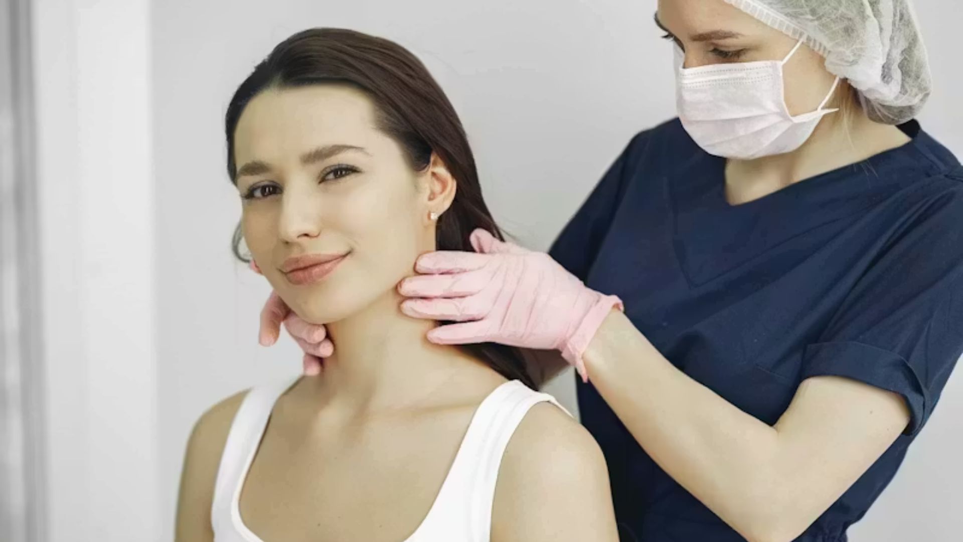 A woman in a white tank top sits calmly as a healthcare professional in scrubs and gloves examines her neck. The setting is clinical and professional.