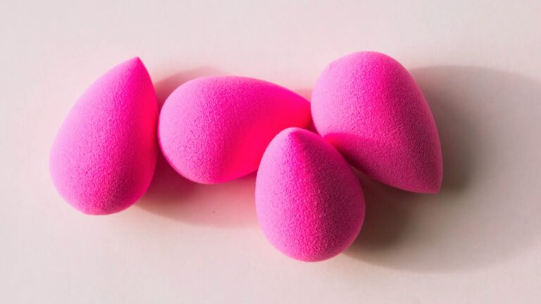 Four bright pink makeup sponges are arranged on a light surface. They have a teardrop shape and a soft texture, conveying a playful and vibrant tone.