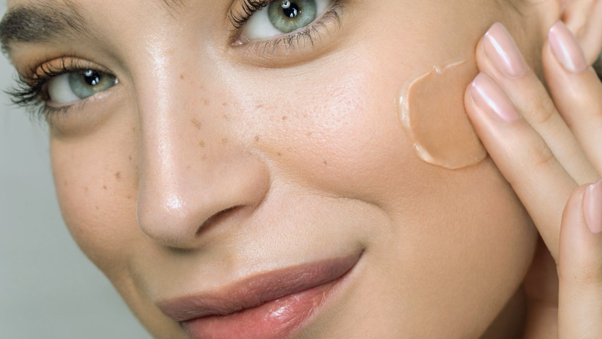 Close-up of a smiling person applying a beige cream to their cheek with their fingertips. The skin appears smooth and radiant, conveying a fresh, healthy look.