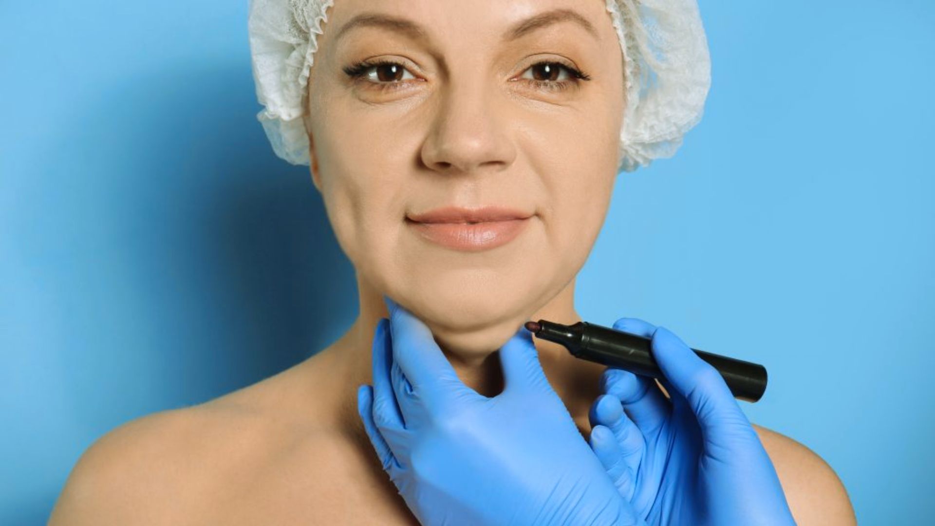 Woman with a neutral expression wearing a hair cap while blue-gloved hands mark her chin with a black marker against a blue background.