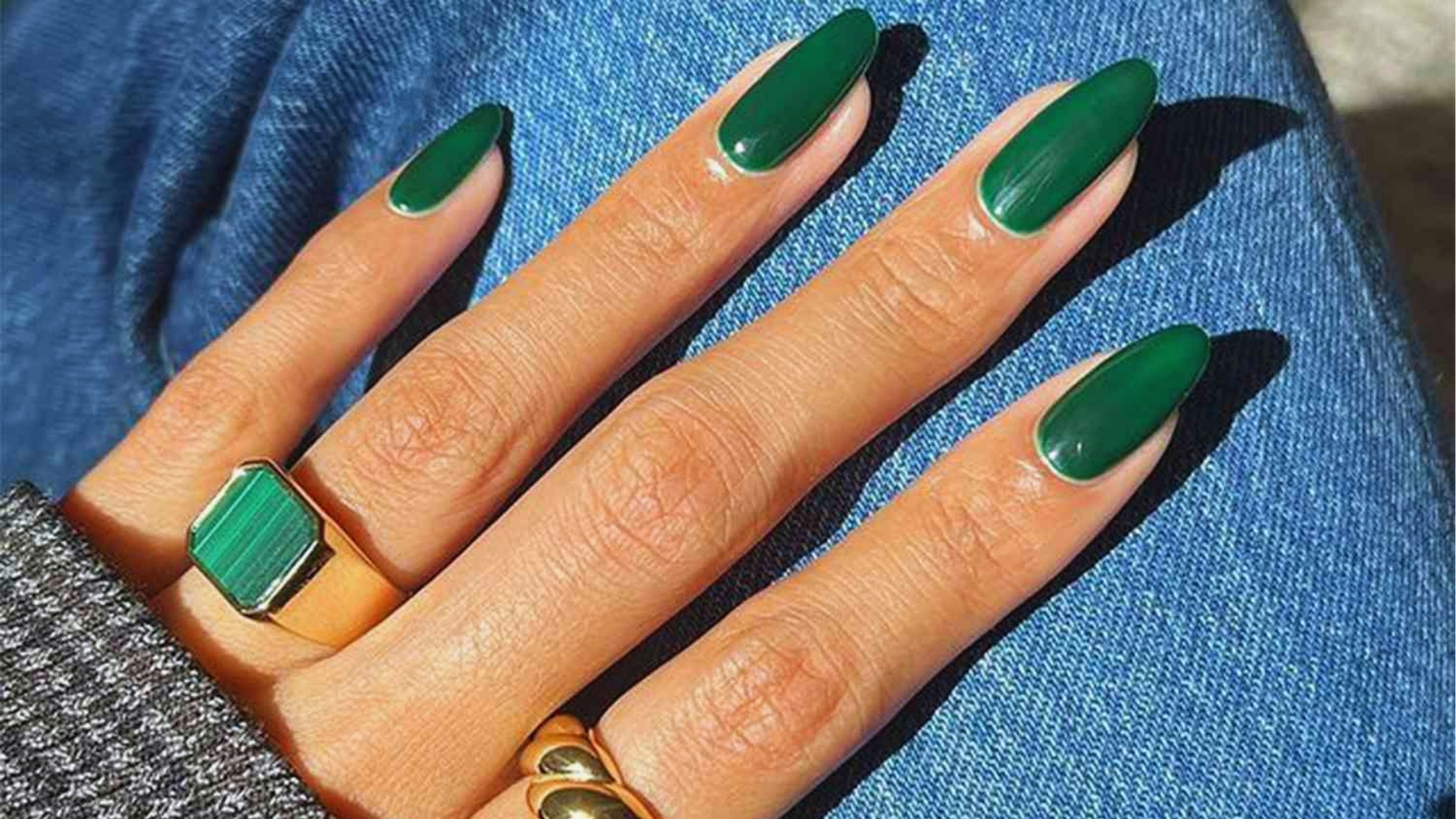 Close-up of a hand with long, almond-shaped nails painted deep green, resting on blue denim. Two gold rings adorn the fingers, adding elegance.