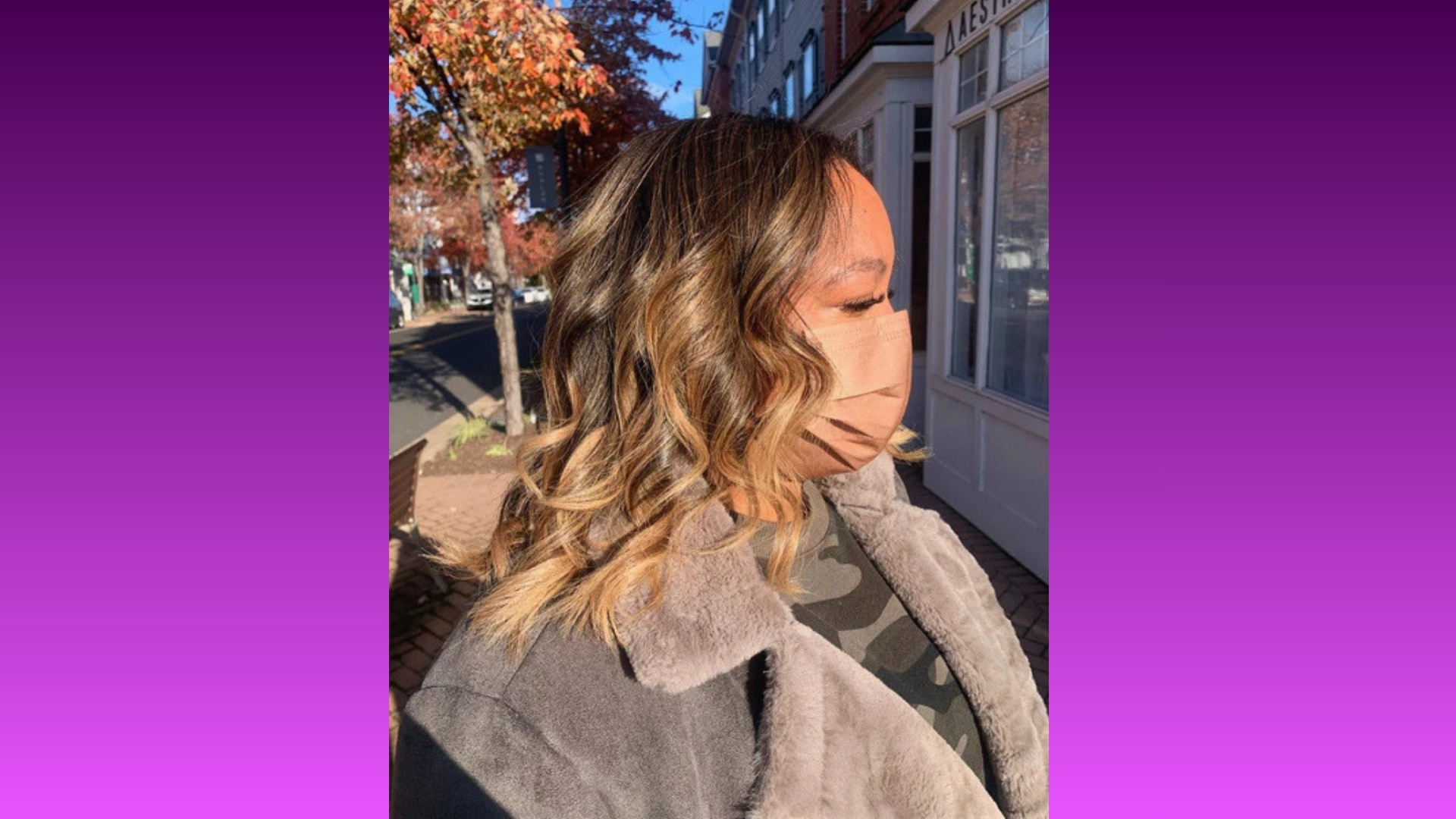 A woman with wavy, highlighted hair wears a face mask and a gray coat, standing on a sunlit street with autumn trees and a storefront in the background.