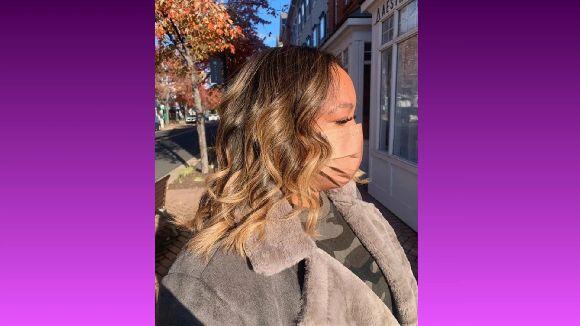 A woman with wavy, highlighted hair wears a face mask and a gray coat, standing on a sunlit street with autumn trees and a storefront in the background.