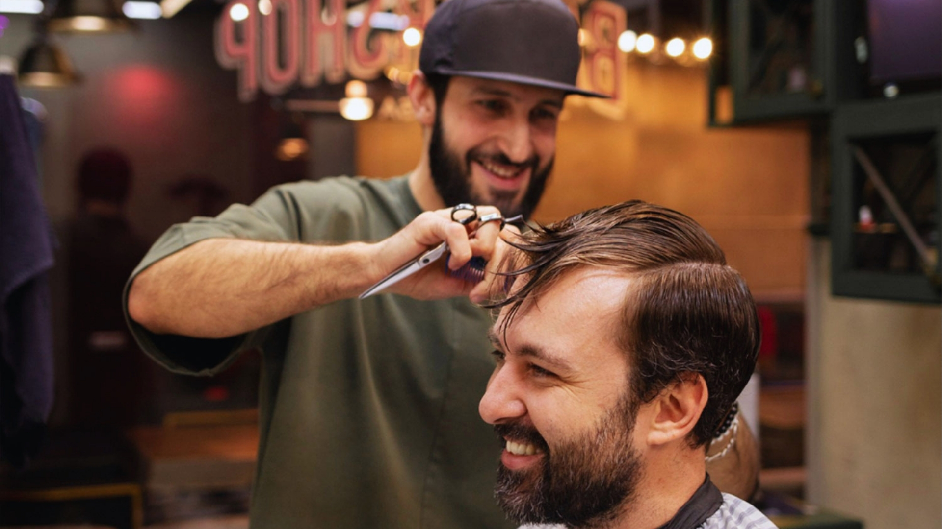A barber with a beard and a cap is cheerfully cutting a client's hair in a cozy barbershop. Both are smiling, creating a friendly, warm atmosphere.