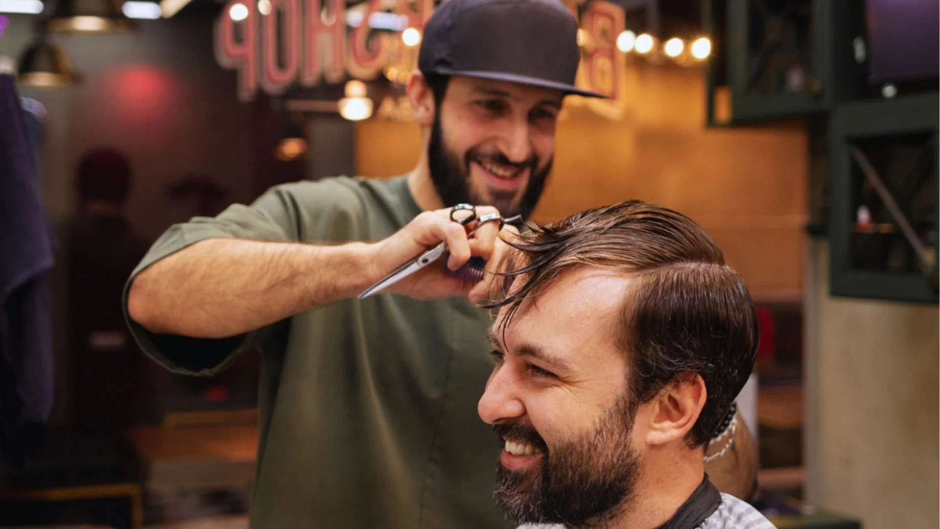 A barber with a beard and a cap is cheerfully cutting a client's hair in a cozy barbershop. Both are smiling, creating a friendly, warm atmosphere.