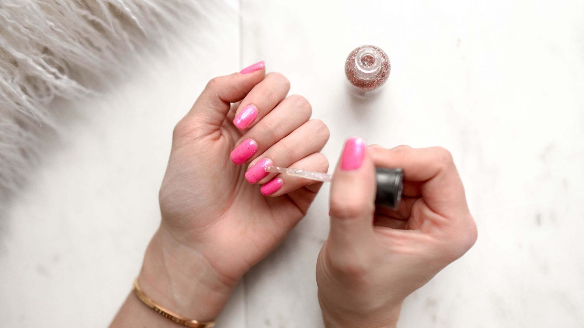 Hands applying pink nail polish on fingernails with a brush. A bottle of polish is nearby on a white surface, creating a serene, self-care vibe.