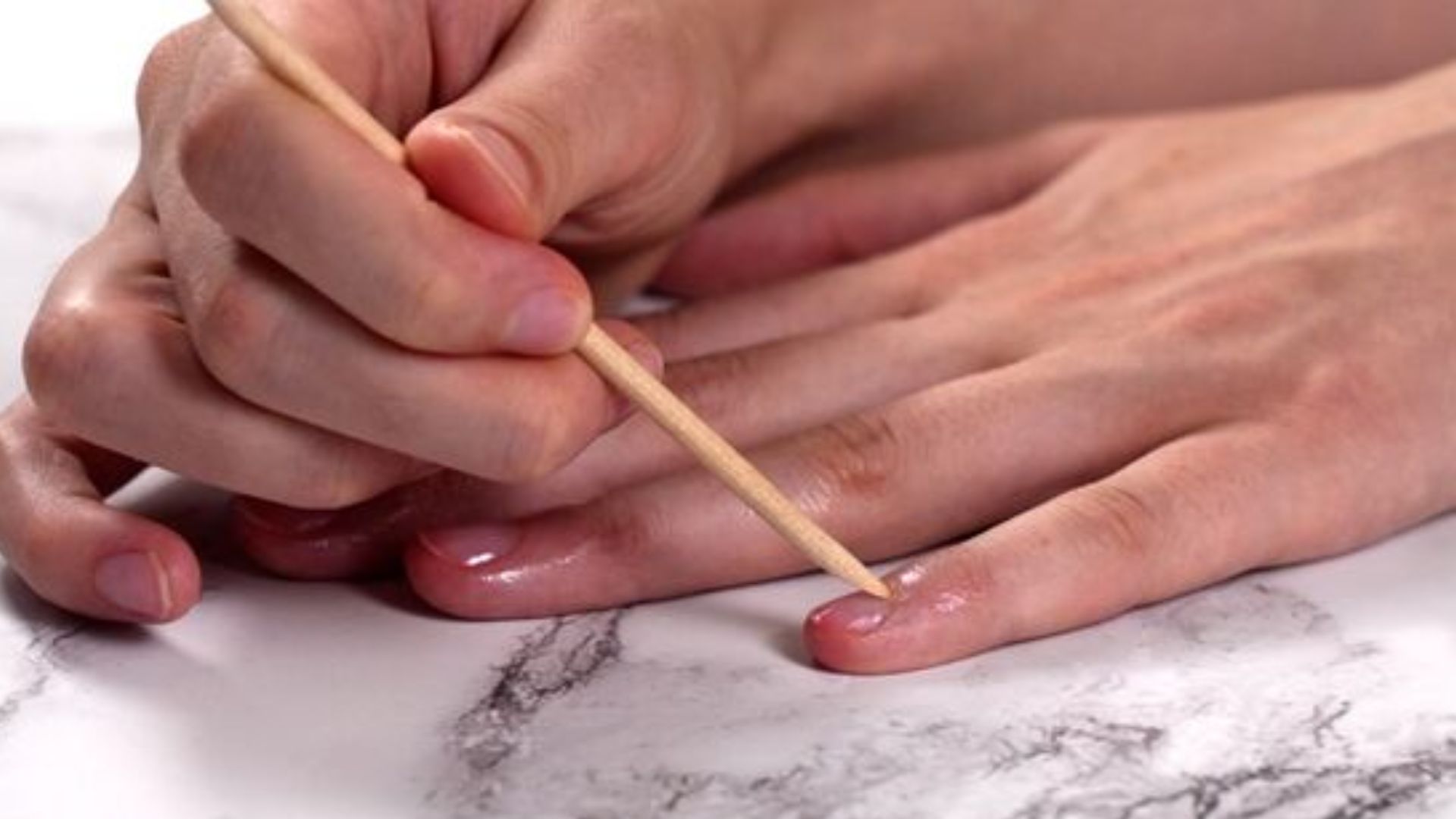 A hand carefully pushes back the cuticles of another hand using a wooden stick on a marble surface, emphasizing a nail care routine and attention to detail.