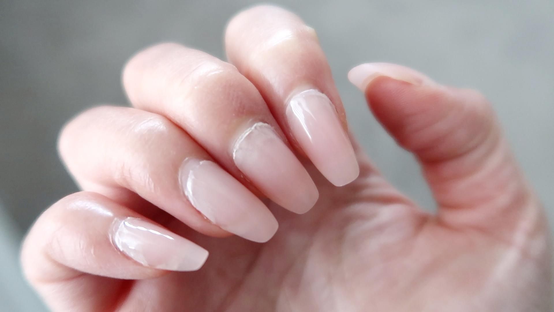 Close-up of a hand with long, almond-shaped nails painted in a glossy, soft pink polish. The background is blurred, highlighting the elegant manicure.