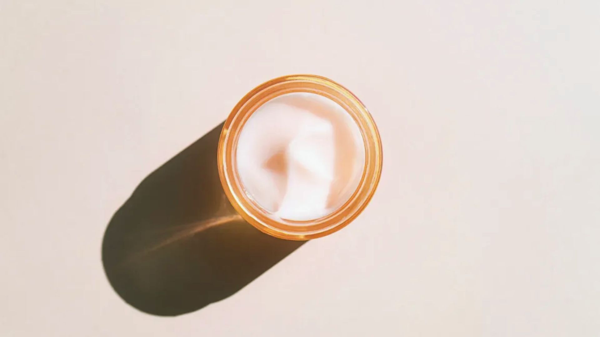A top-view of an open jar of cream on a beige surface. The jar casts a long shadow to the left, creating a minimalist and calm composition.