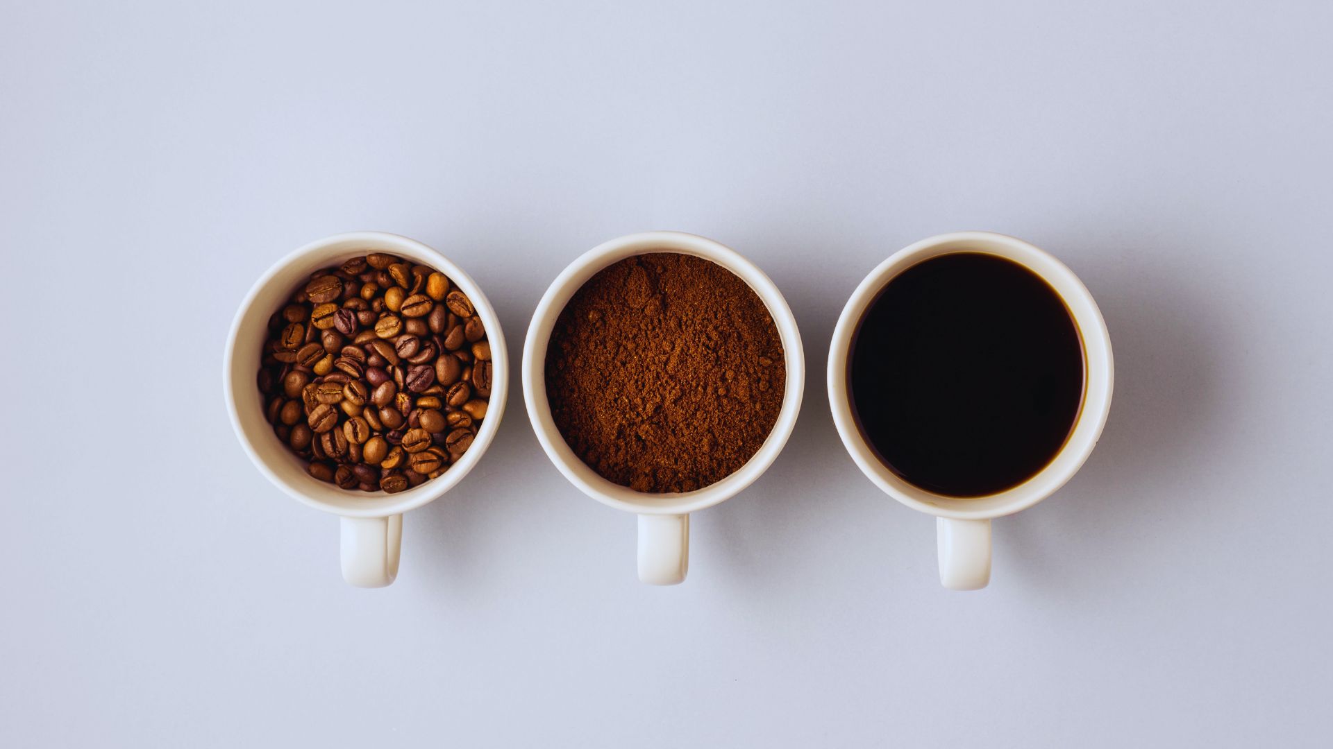Three white cups in a row on a white background; from left: filled with coffee beans, ground coffee, and black coffee, illustrating coffee stages.