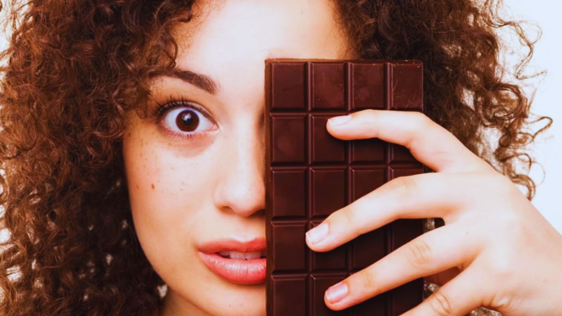A person with curly hair playfully holds a large chocolate bar over one eye, displaying a surprised expression. The image conveys a fun, whimsical tone.