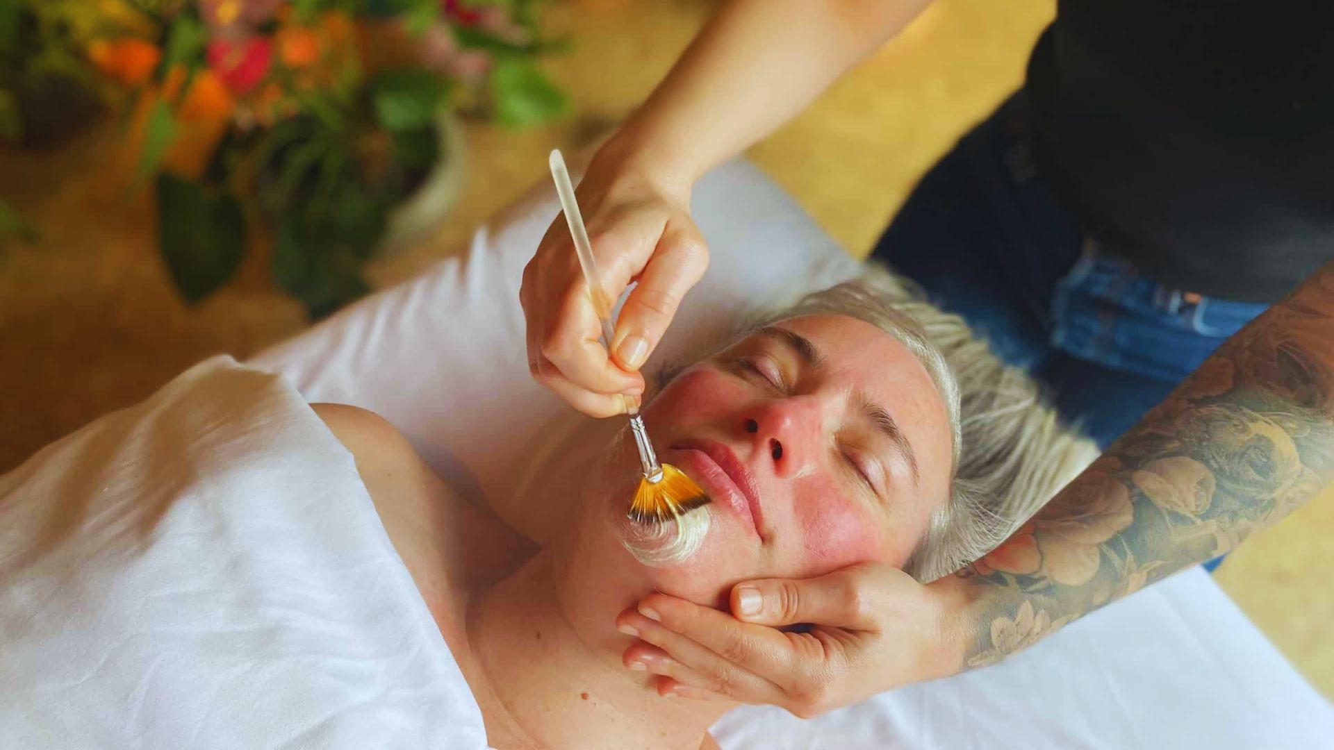 A person receives a relaxing facial treatment with a brush while lying on a white-covered table. The setting is calm, with blurred greenery in the background.