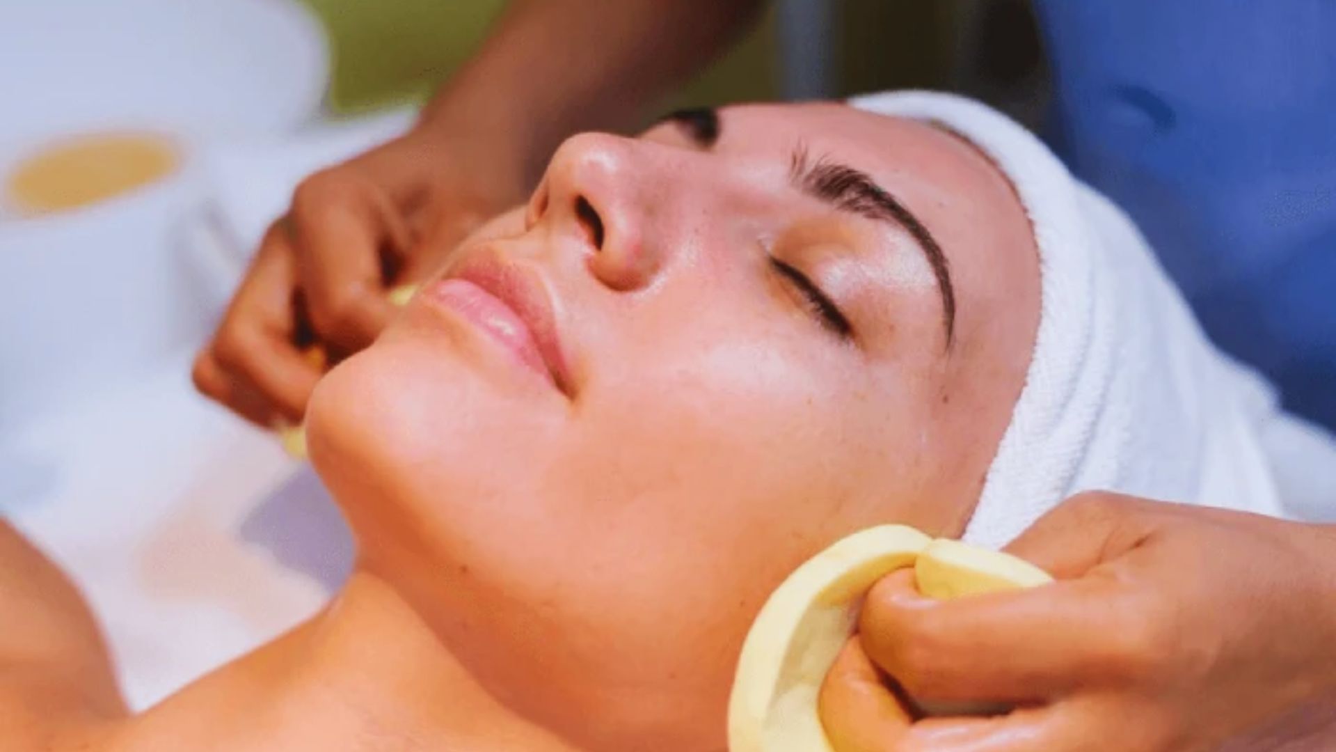 Woman receiving a facial treatment in a spa setting. Her eyes are closed, face relaxed, with a towel wrapped around her head. Hands gently cleanse her skin.