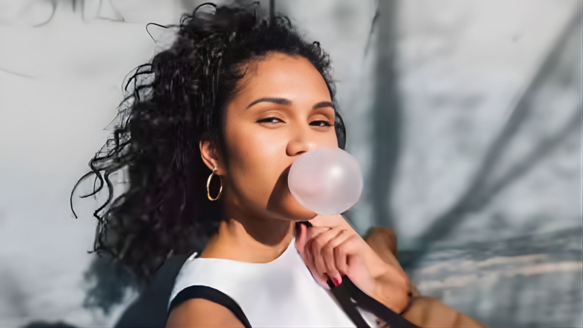 A woman with curly hair and hoop earrings blows a bubblegum bubble, looking confident. She wears a sleeveless top, with shadows on a light background.
