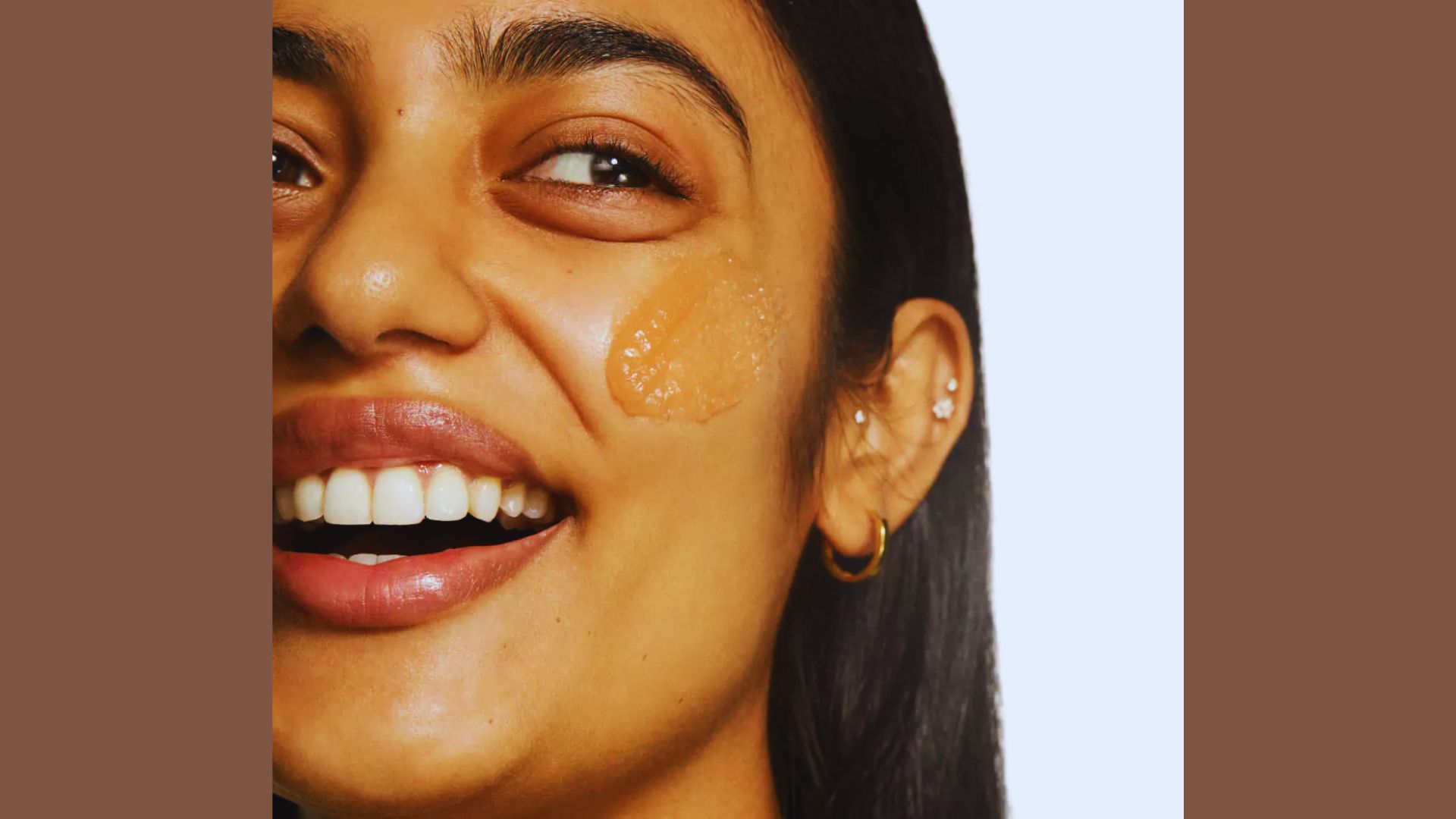 Close-up of a smiling person with smooth skin and a patch of skincare product on their cheek. They have earrings and are set against a light background.