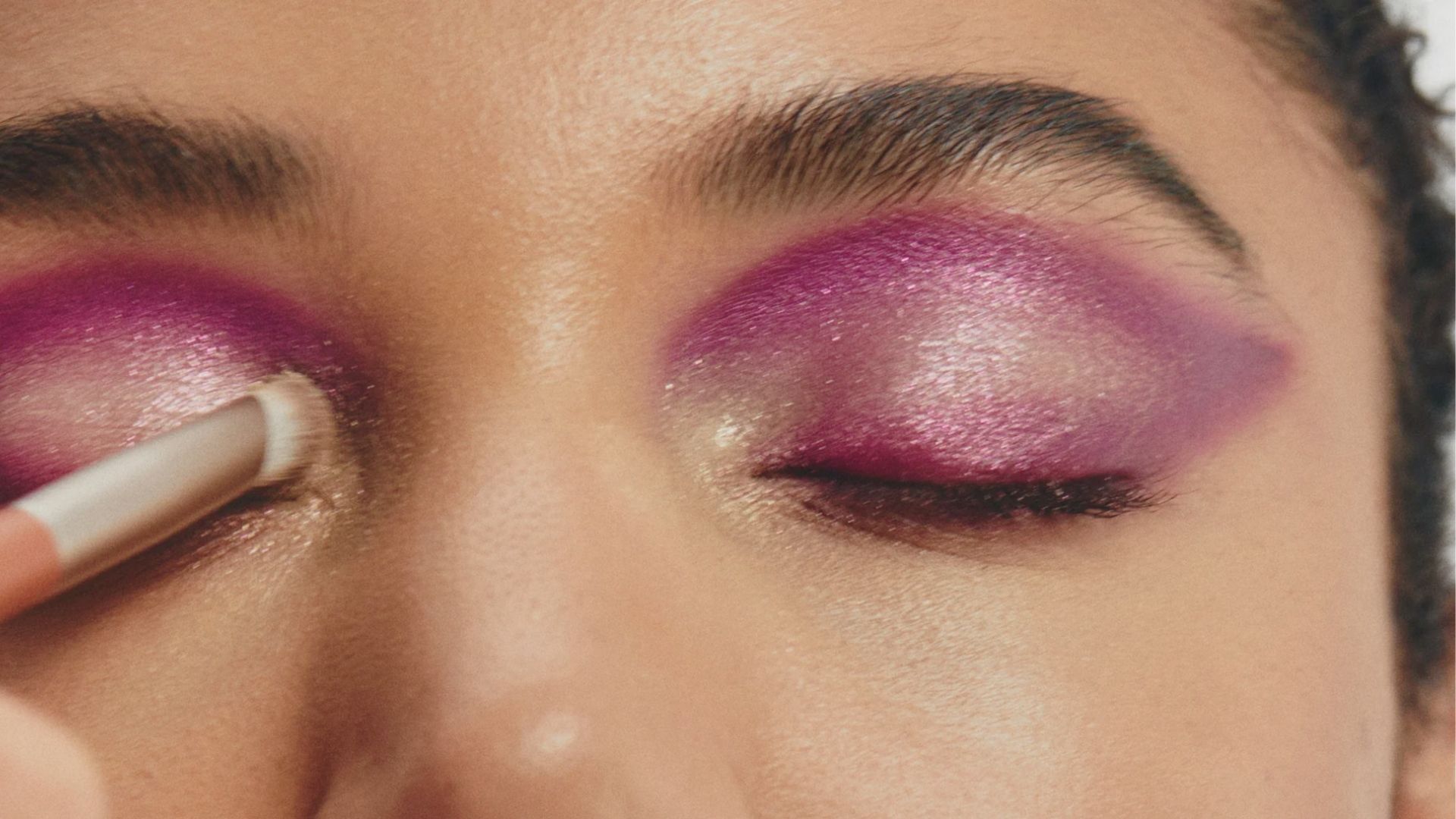 Close-up of an eye with vibrant pink and shimmering eyeshadow being applied. The bold makeup creates a striking and glamorous look.