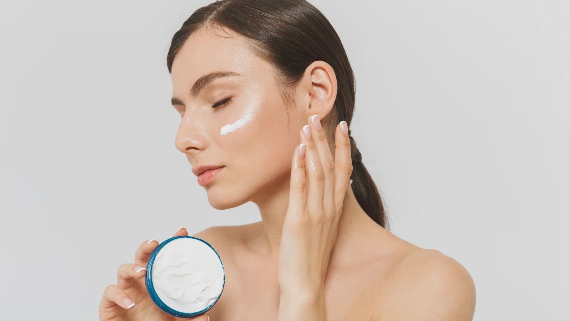 A woman with closed eyes applies white cream to her cheek, holding a jar. She has a calm expression and a smooth, neutral background.