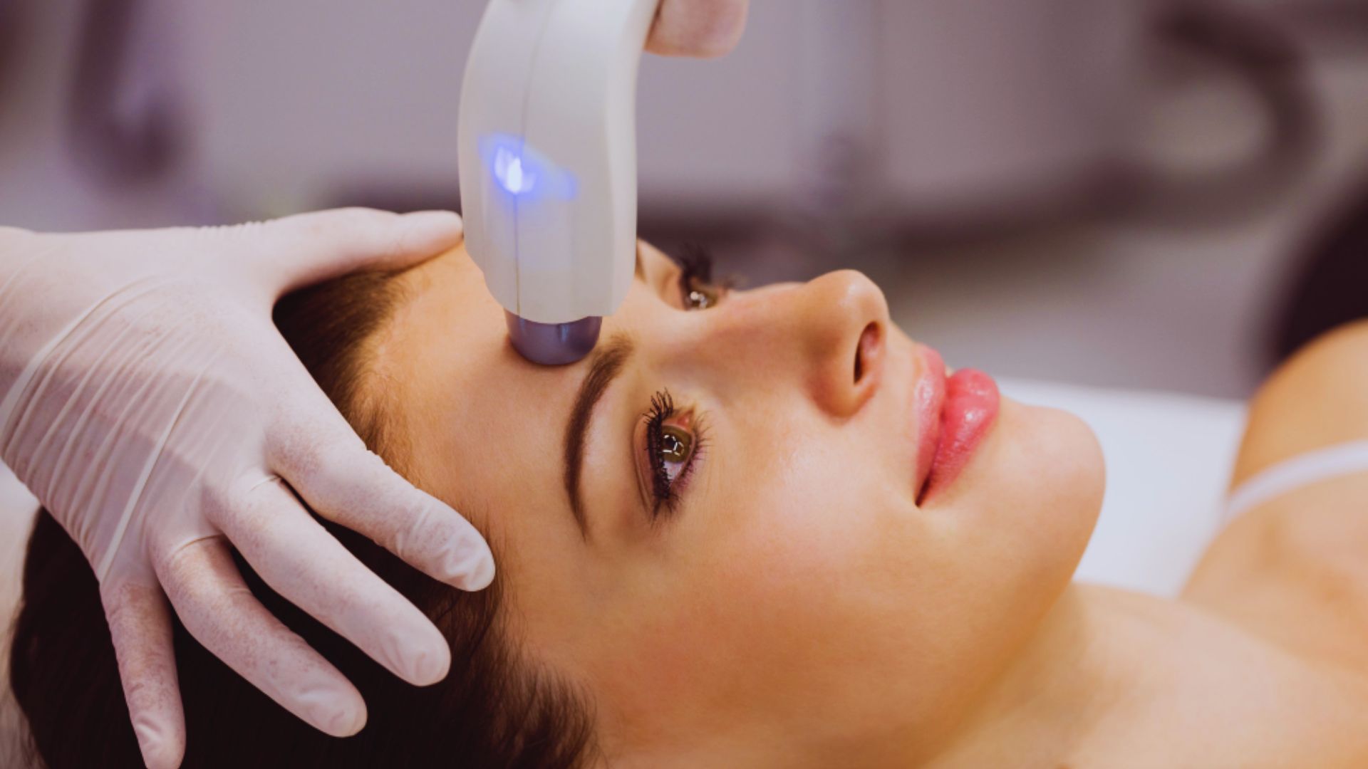 A woman receives a beauty treatment, with a device applied to her forehead. She looks relaxed, while a gloved hand gently holds her face.