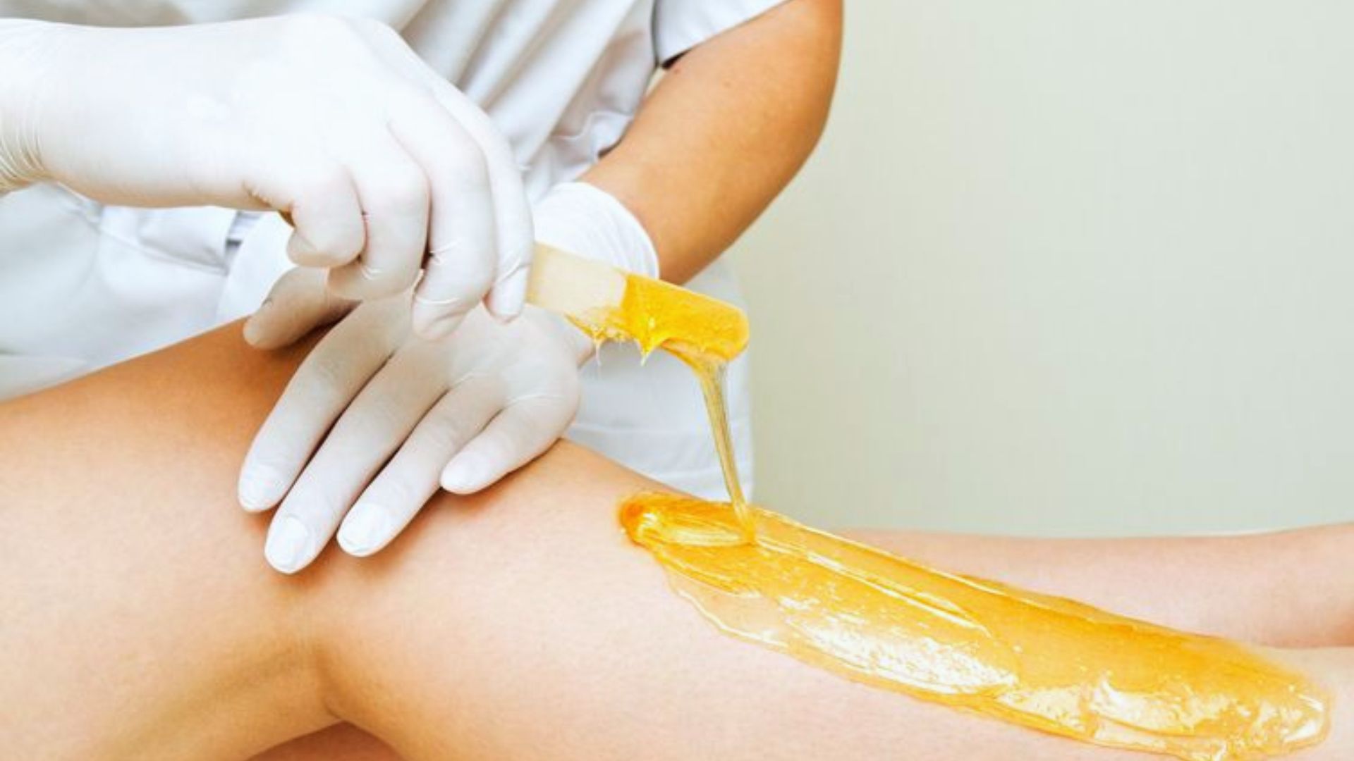 A person applies warm honey-colored wax on a leg with a spatula. They wear white gloves, suggesting a clean, professional beauty treatment setting.