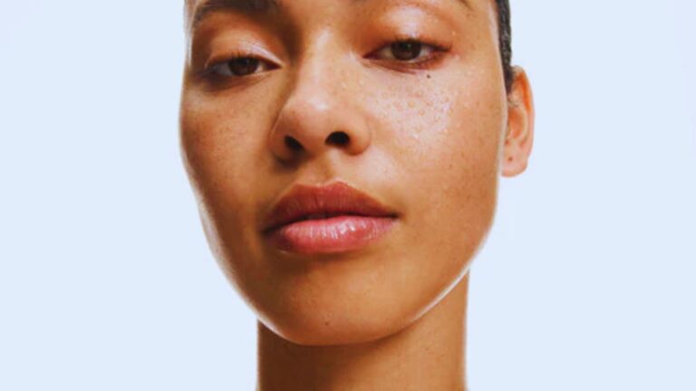 Close-up of a person with freckles, gazing forward with a serene expression. The lighting highlights smooth skin and natural features against a pale blue background.