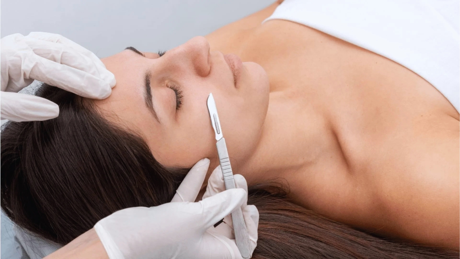 A woman lies calmly with eyes closed, receiving a dermaplaning treatment. Gloved hands hold a small scalpel near her cheek, focusing on exfoliation.